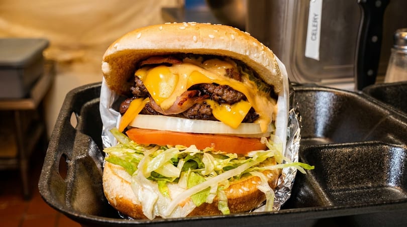 A double cheeseburger with bacon, cheese, lettuce, tomato, and onion in a black takeout container.