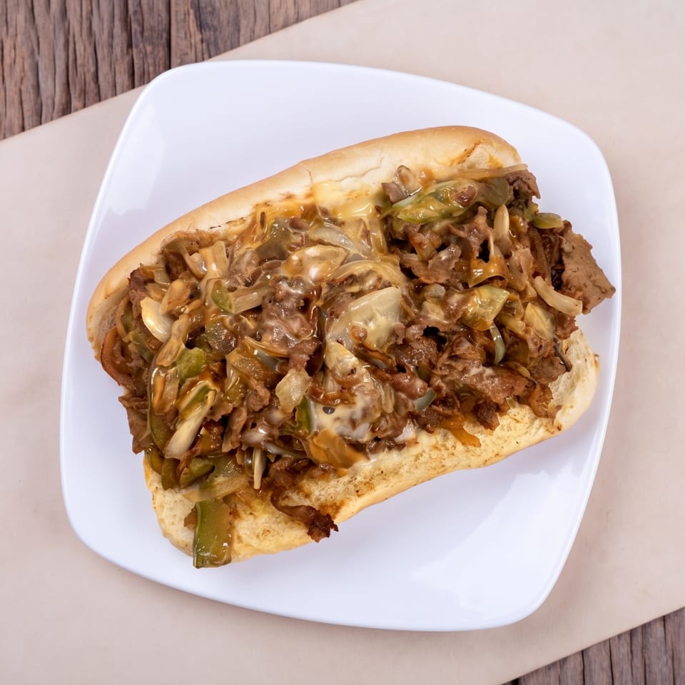 Hearty cheesesteak sandwich with melted cheese, steak, onions, and peppers on a white plate.