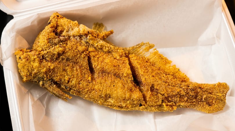 Whole fried fish, golden brown and crispy, in a white takeout container.