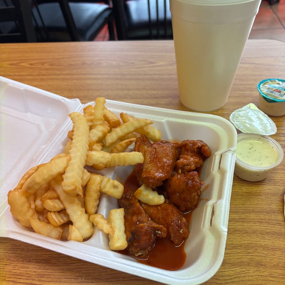 A takeout container with buffalo wings, crinkle-cut fries, a drink, and dipping sauces on a wooden table.