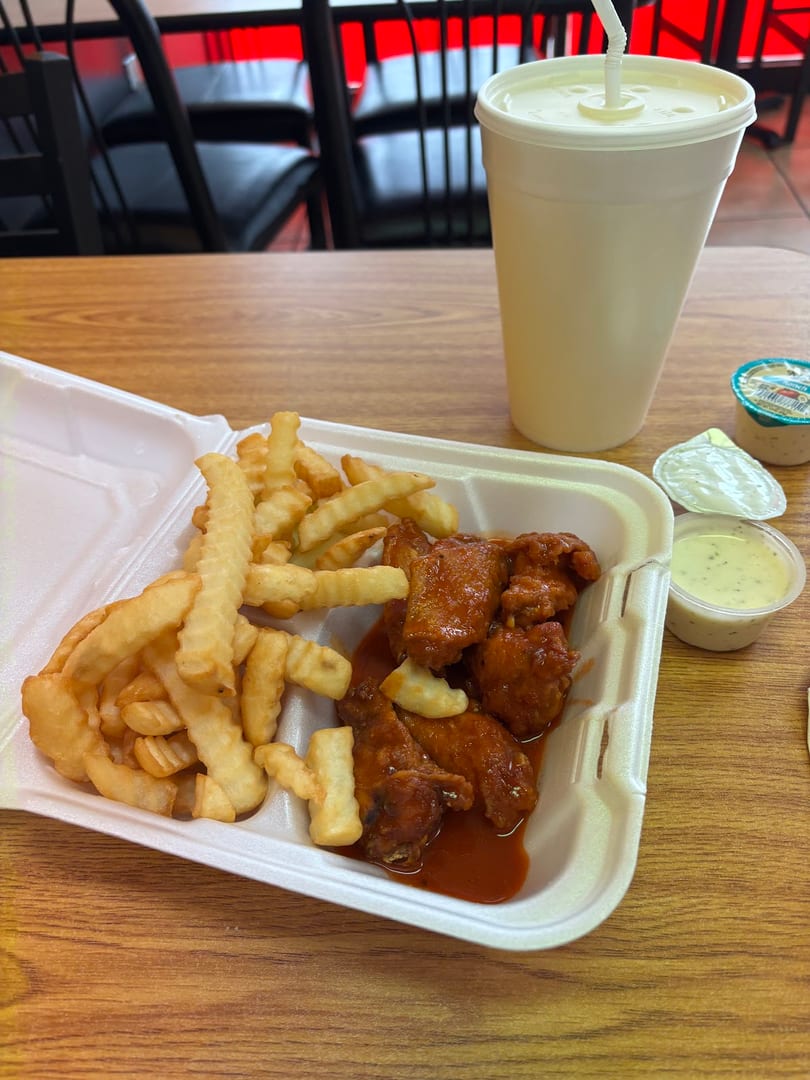 A takeout container with buffalo wings, crinkle-cut fries, a drink, and dipping sauces on a wooden table.