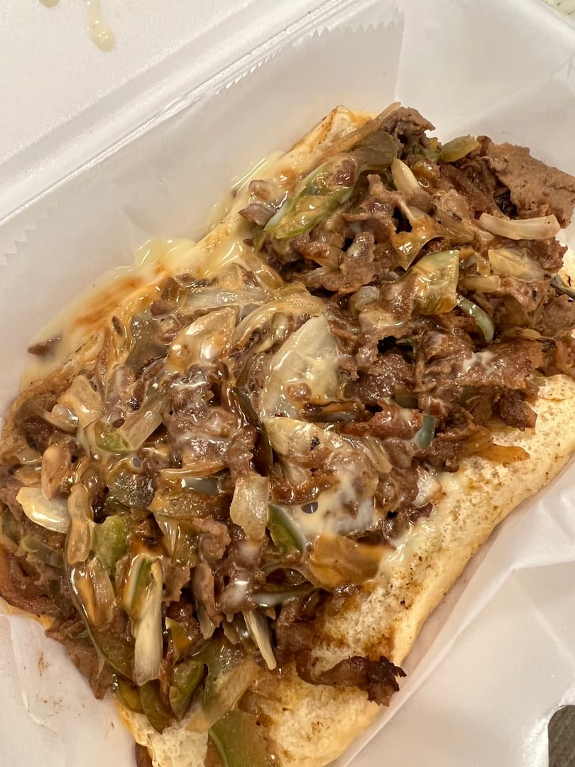 Cheesesteak sandwich with steak, melted cheese, onions, and green peppers in a white container.